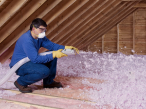 Technician blowing pink loose-fill insulation into an unfinished attic.