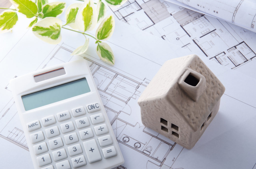 White figurine of a house next to a white calculator, on top of a blue print.